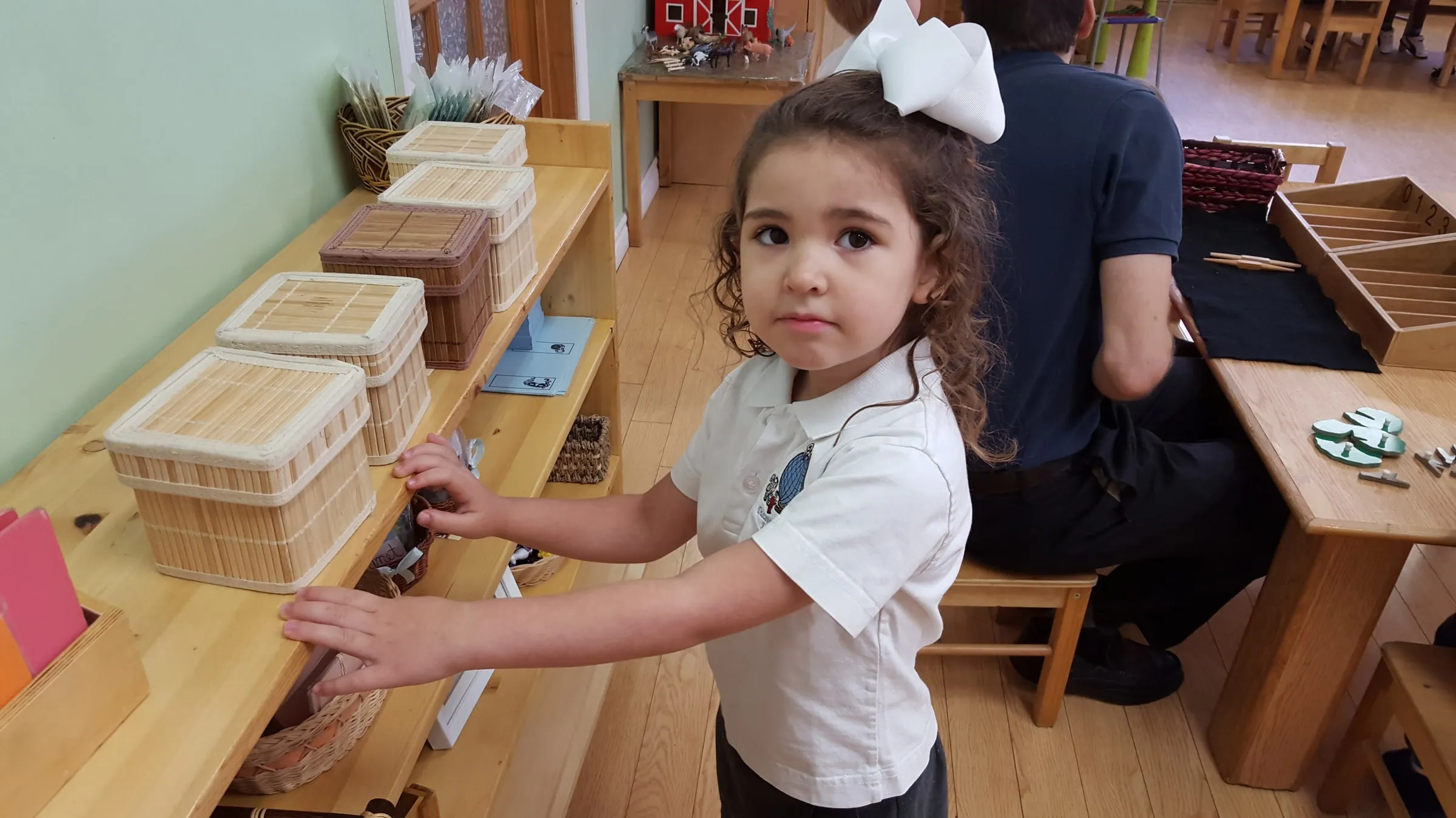A young girl in school uniform choosing a basket of work from a low classroom shelf