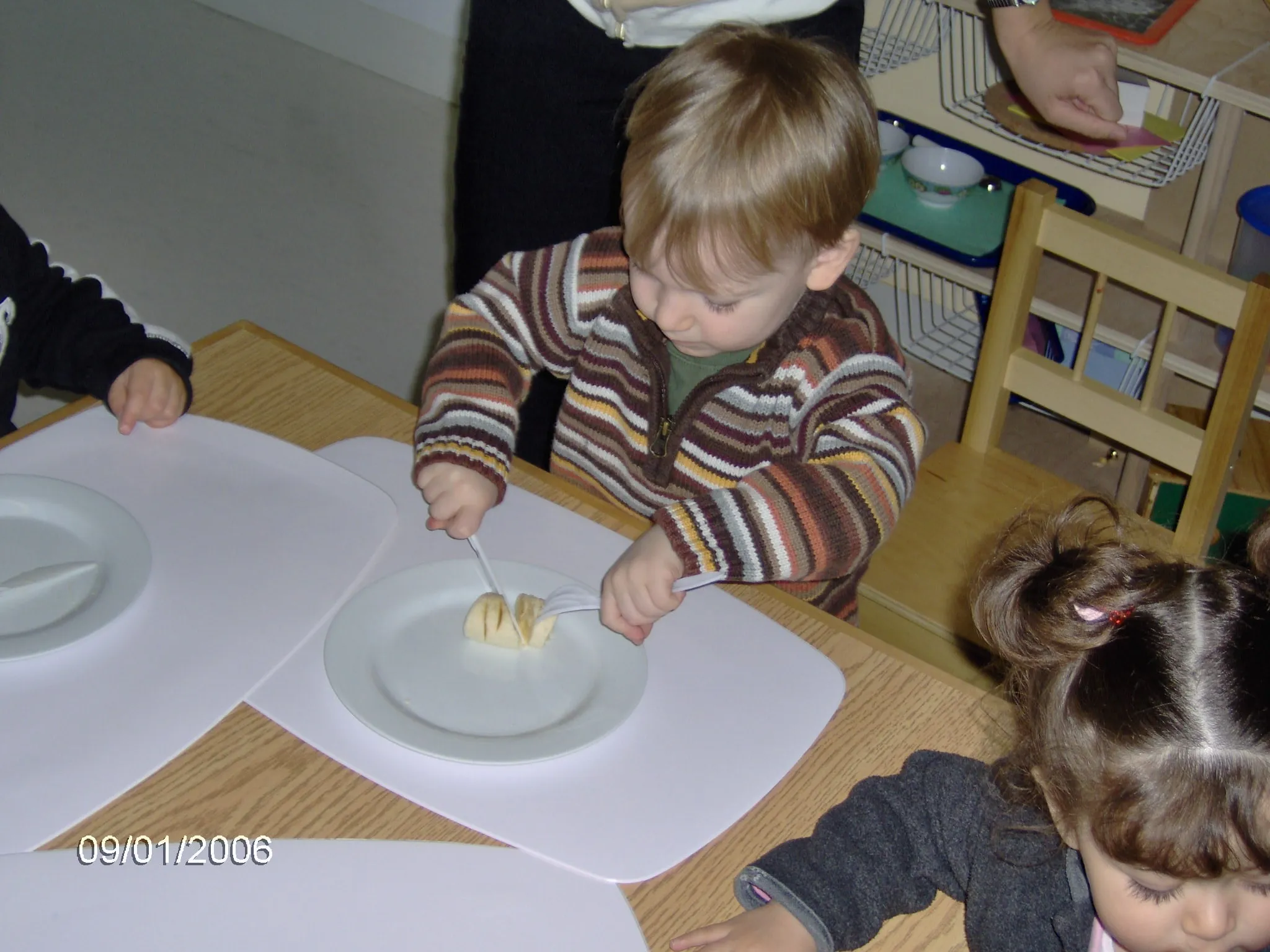 A toddler in a striped jumper carefully slicing a banana with a table knife, watched by a teacher