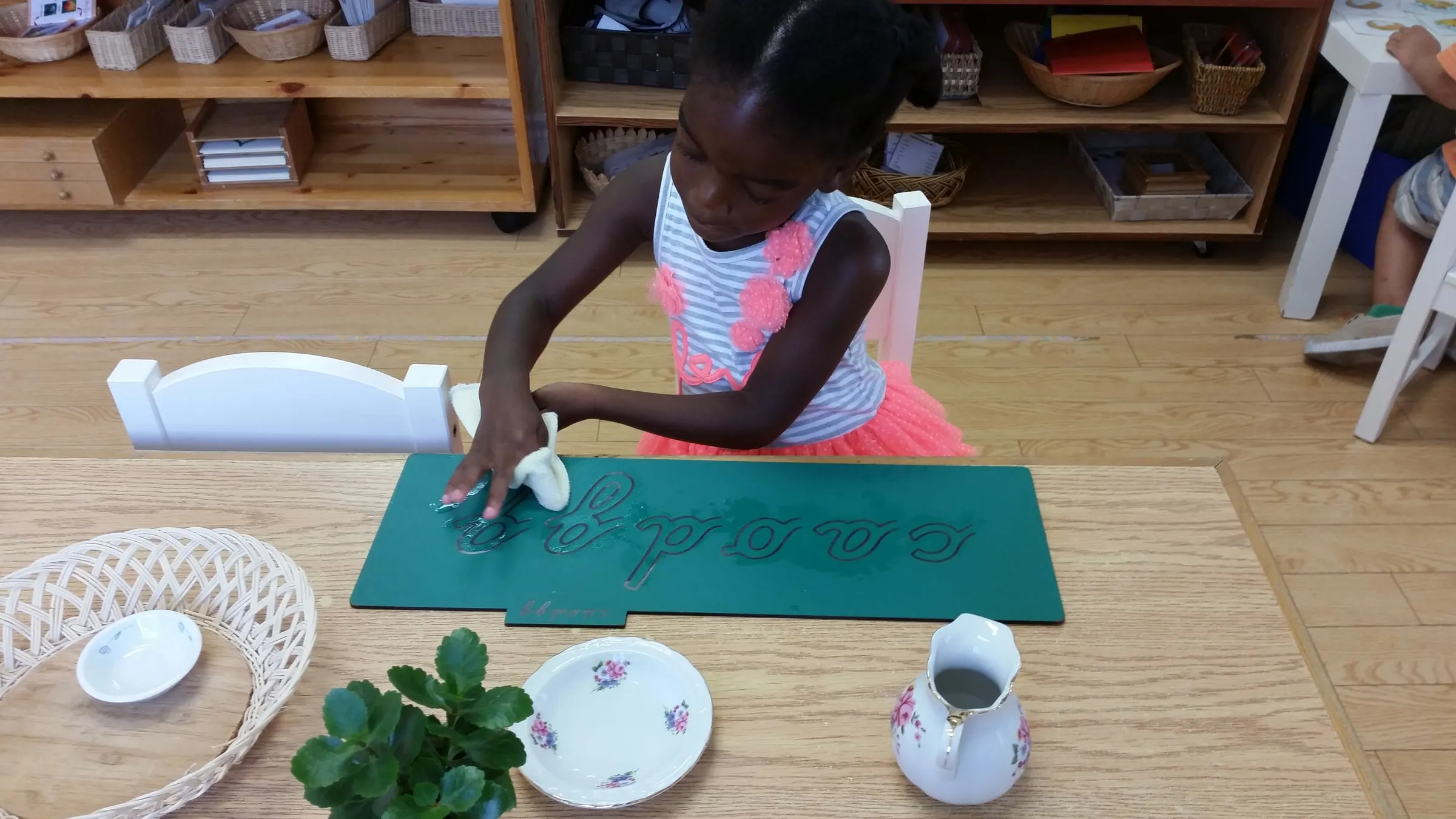 A young girl tracing cursive letters on a green board with a damp cloth