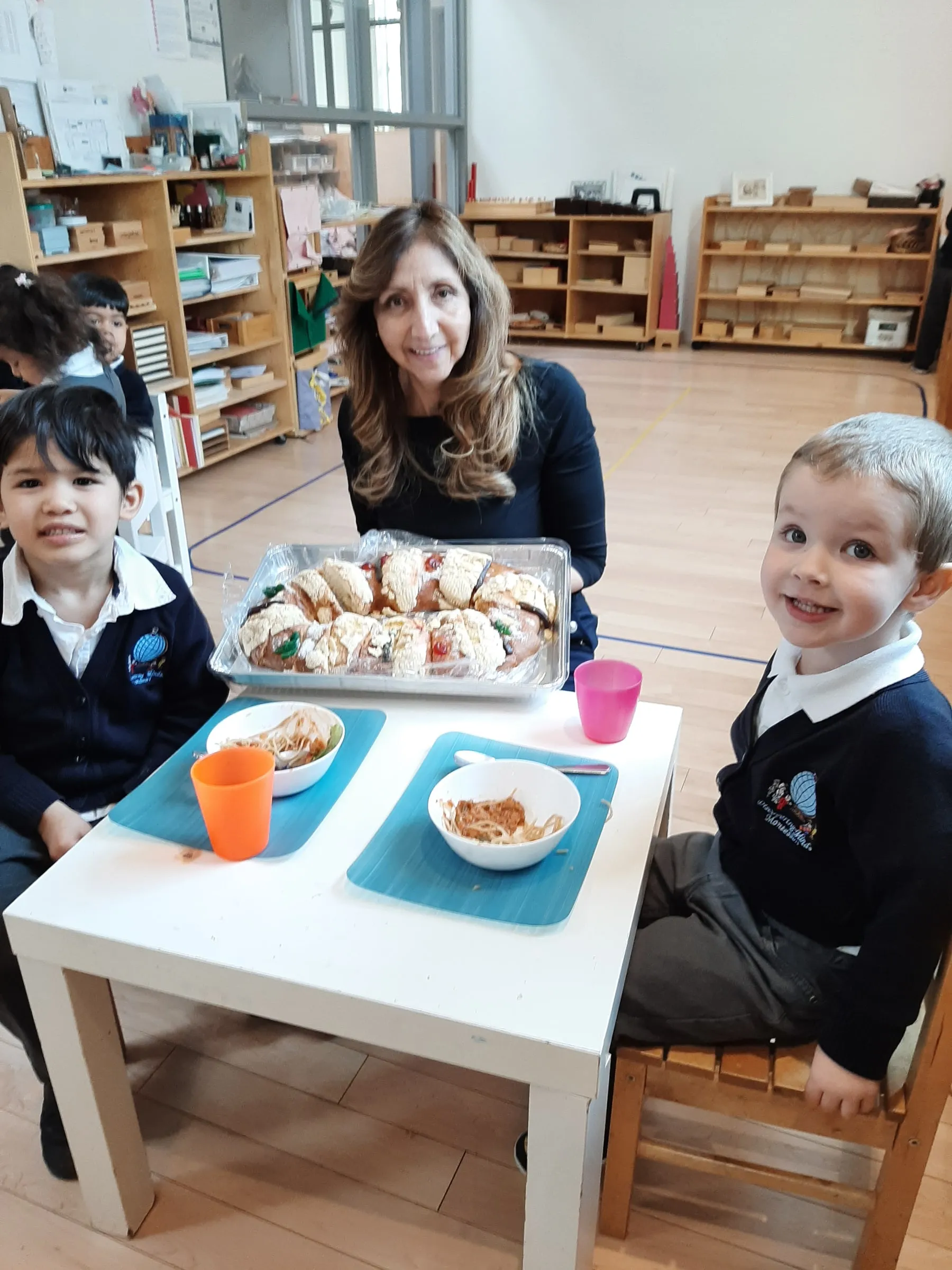 A teacher sharing lunch at a low table with two young children in the classroom